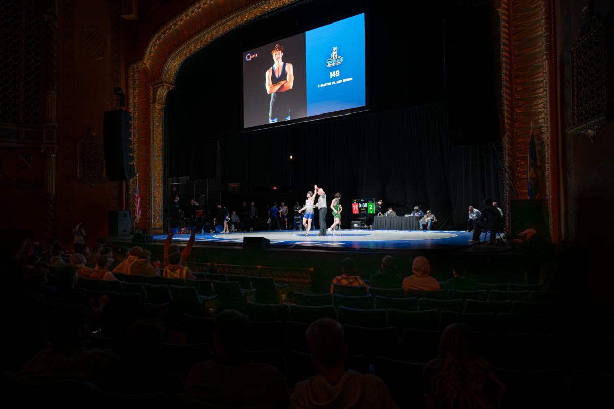 Wide shot of the wrestling stage inside a theater, showing the mat, scoreboard, teams, and audience seating.