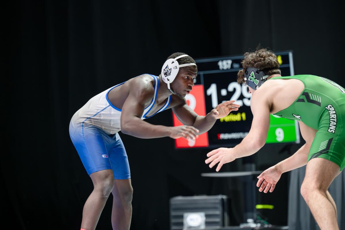 Two wrestlers face each other in a neutral stance on the mat, hands extended and a digital scoreboard visible in the background.
