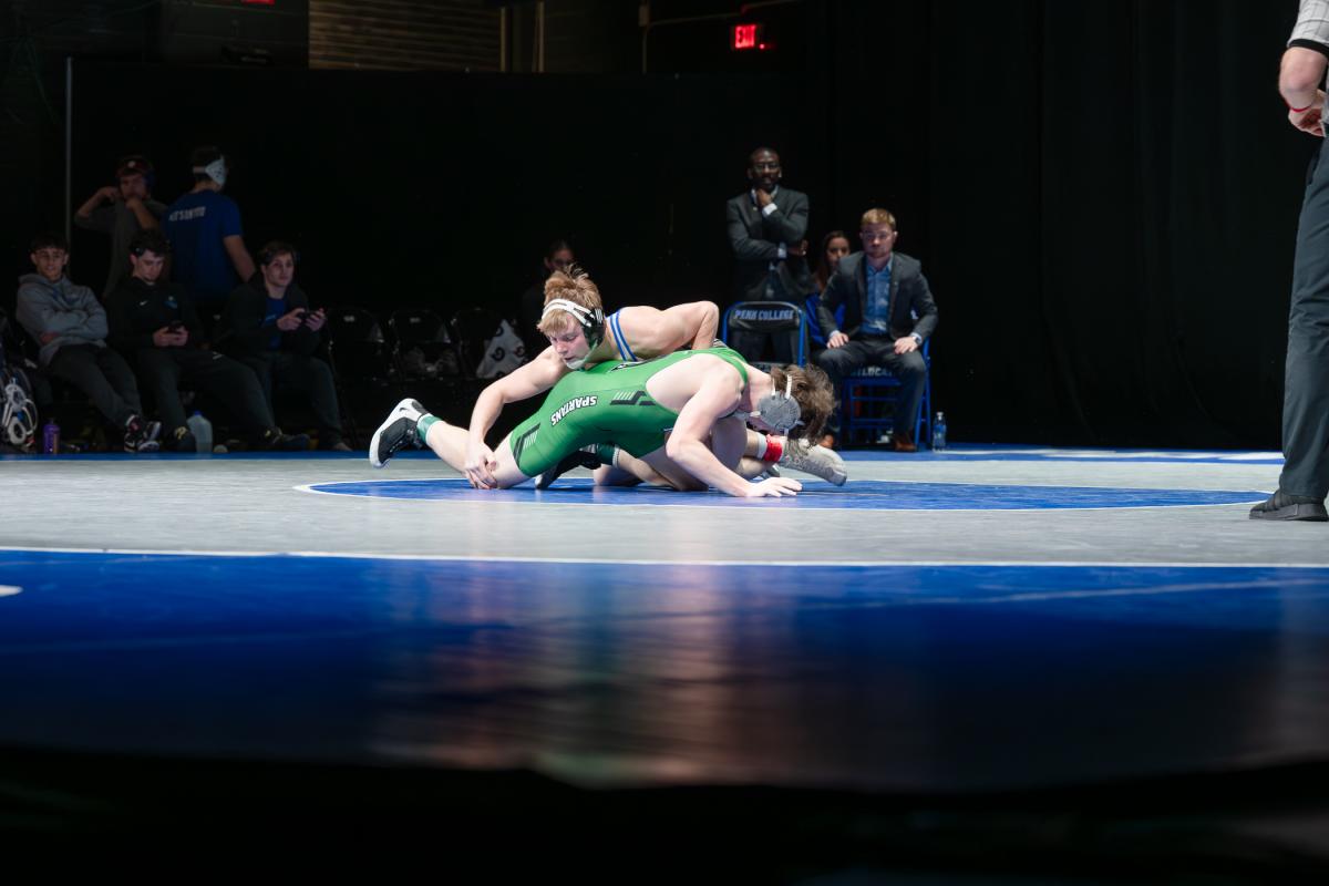 Two collegiate wrestlers compete on a blue mat during a live match, with one athlete controlling the other on the mat as officials and spectators watch.