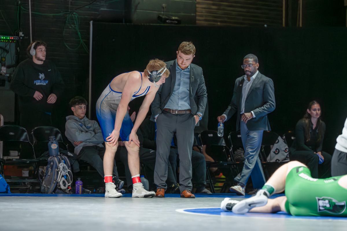 A wrestler bends forward at the edge of the mat while coaches and staff stand nearby during a break in the match.