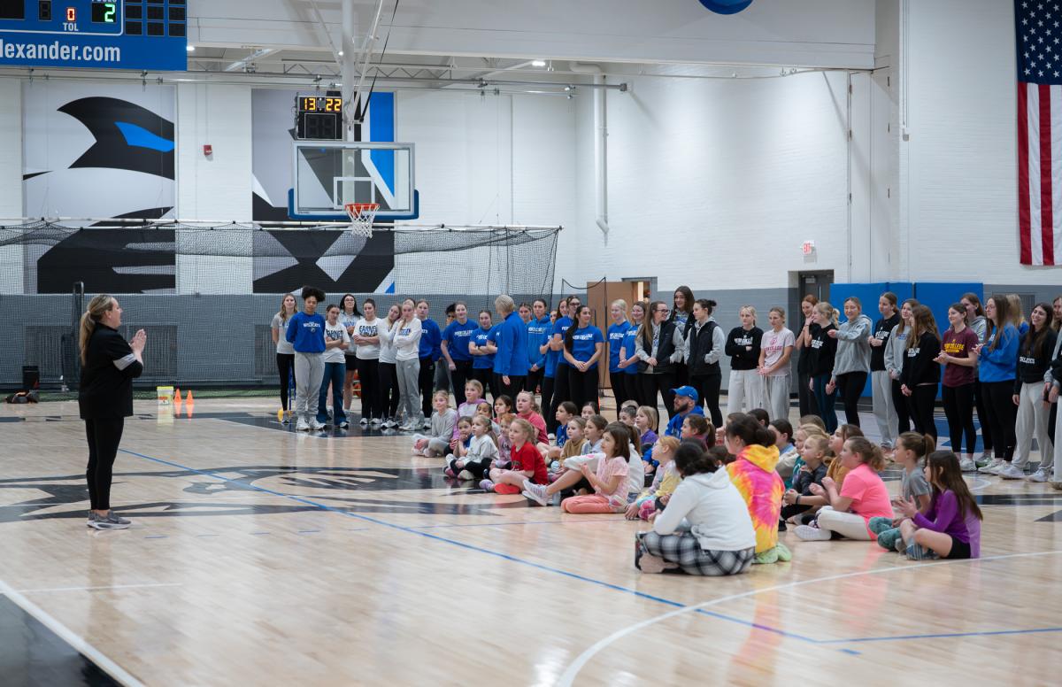 A group of girls sits on a gym floor with student-athletes standing in a group behind them, while a woman stands in front of them, speaking.