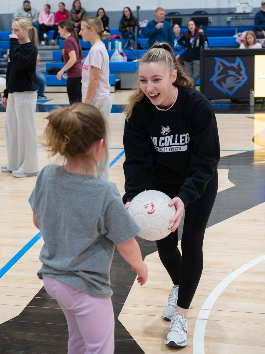 A female college athlete smiles widely while holding a soccer ball and interacting with a girl.