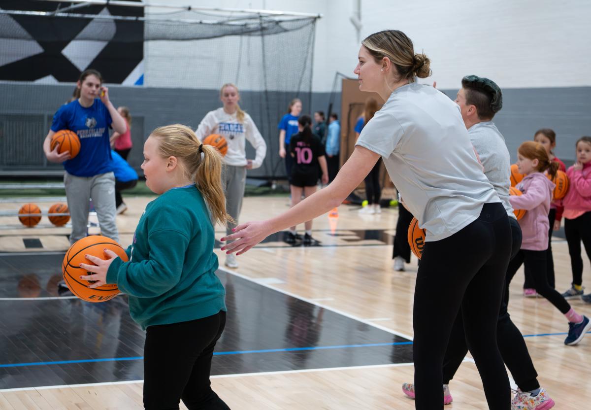 A child holds a basketball while a female college athlete encourages her.