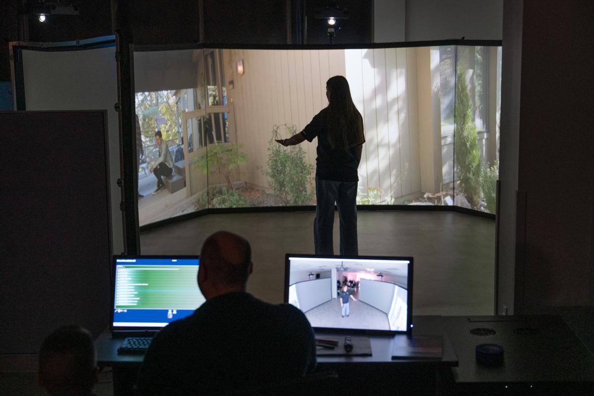 While one person sits behind a computer monitor, another stands and talks with an actor on a video screen.