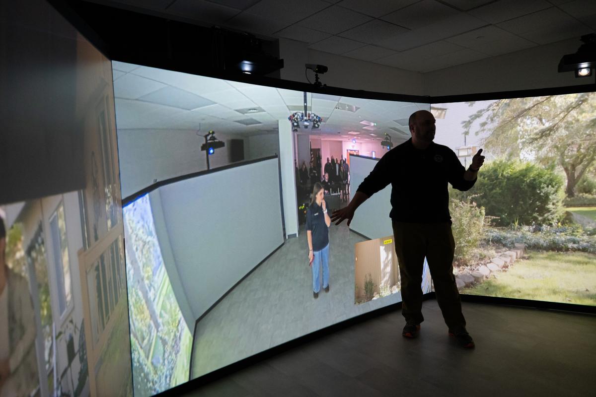 A person stands and speaks while surrounded by video screens.