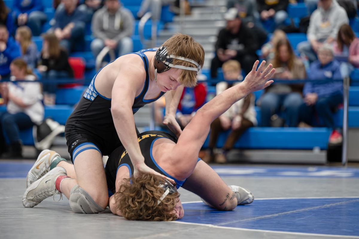 One wrestler maintains control on the mat while positioned above an opponent who is reaching outward with one arm. Spectators sit in bleachers in the background.