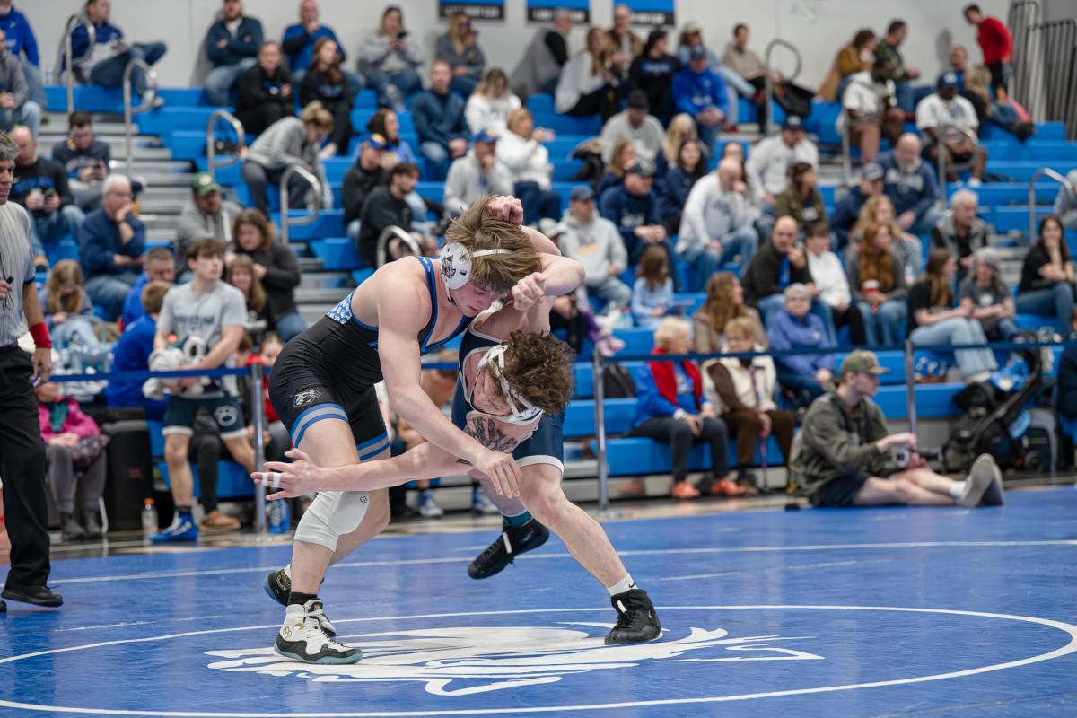 Two wrestlers in a bout in the center of a blue wrestling mat in Penn College's Bardo Gym. Bleachers in the background are full of spectators.