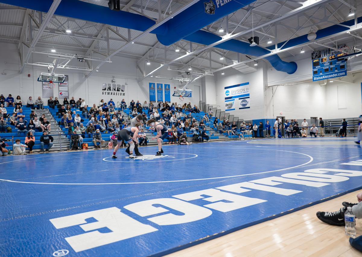 Wide view of two wrestlers at the center of a mat in Penn College's Bardo Gym, showing full bleachers.