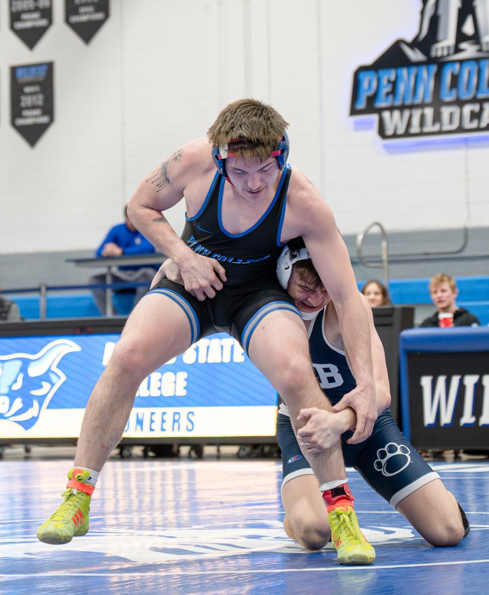 Two wrestlers compete during a match on a blue mat, with one athlete standing and attempting to maintain balance while the other athlete holds a leg from a lower position. Penn College wrestling signage is visible in the background.