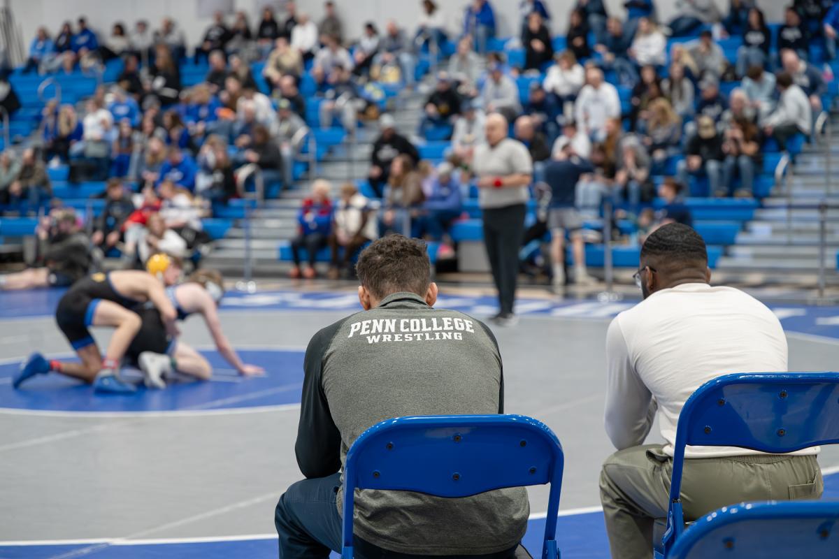 Two individuals seated in blue chairs observe a wrestling match in progress on a gymnasium floor. One wears apparel labeled ‘Penn College Wrestling.’ A crowd of spectators fills the bleachers behind them.