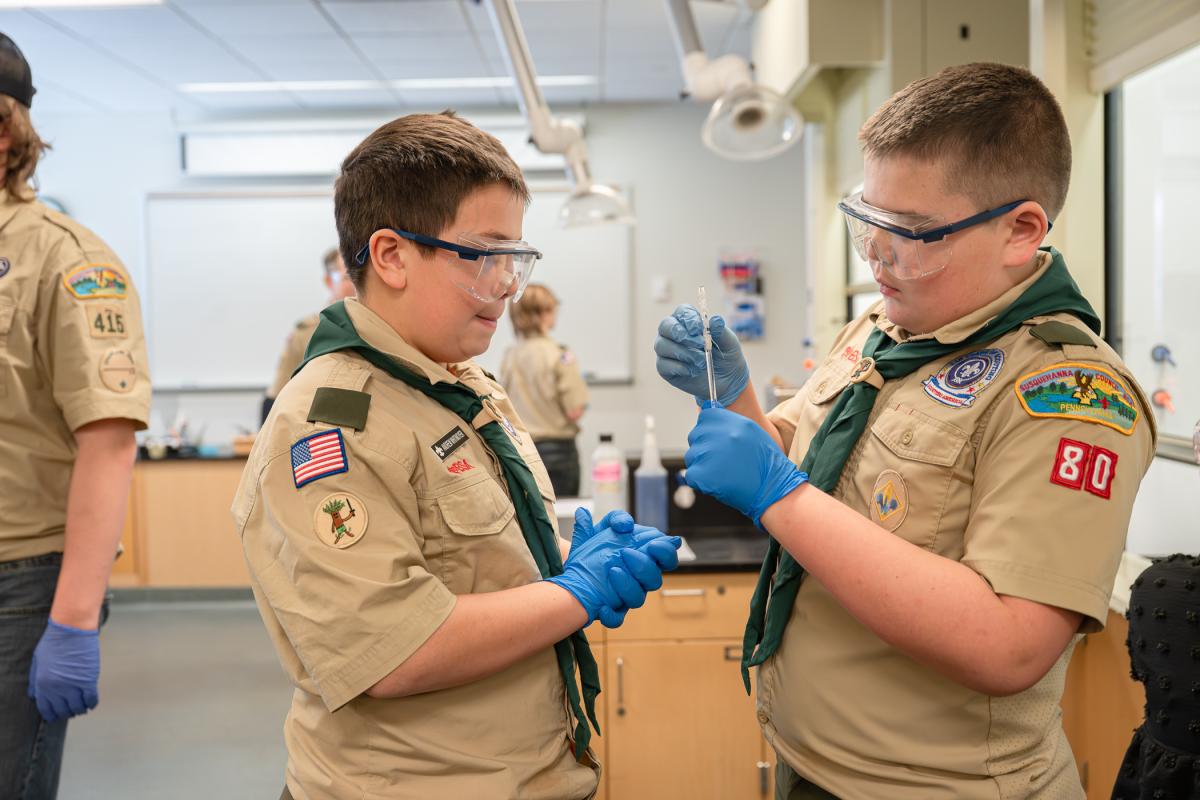 Two boys in Scouting America uniforms wear safety goggles and rubber gloves as one inspects a substance on the end of a glass stirring rod.