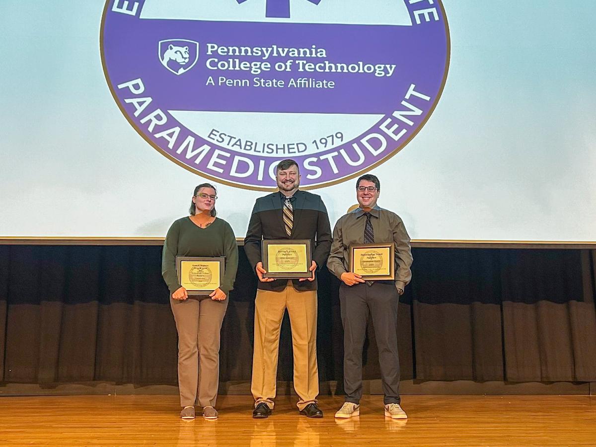 Three people stand on a stage. Each is holding a framed certificate.