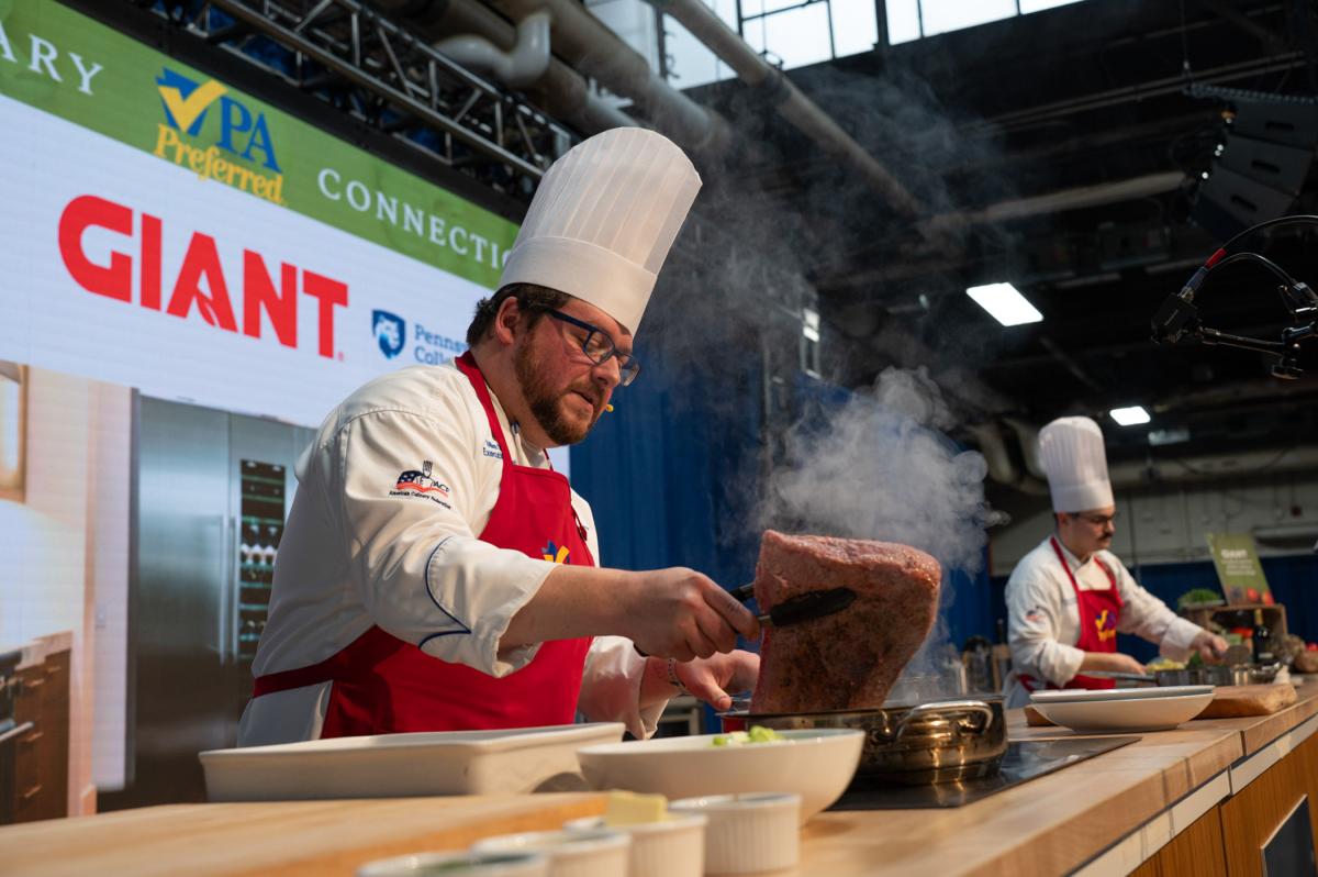 Chef slicing and searing a large cut of meat on a cooking station during a live event.