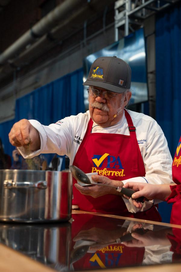 Close-up of a chef seasoning food in a pot while another person observes.