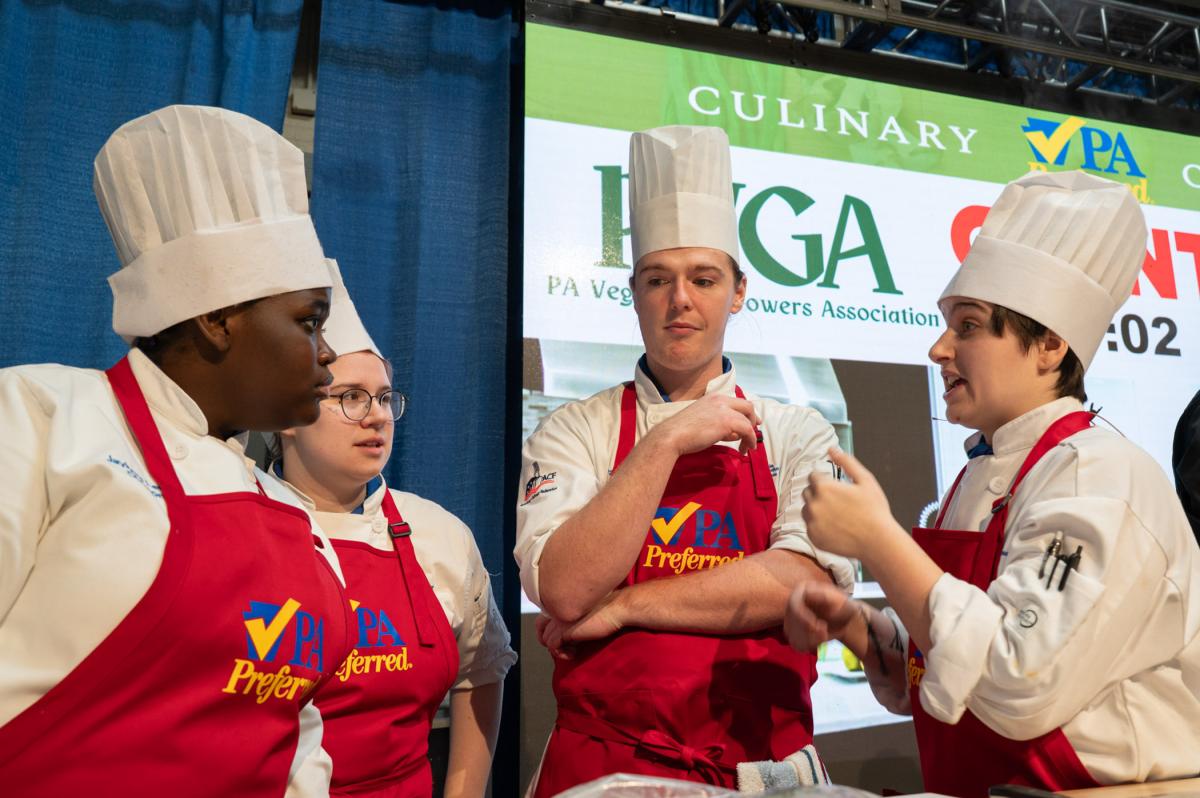 Three chefs in aprons discussing food preparation at a competition station.