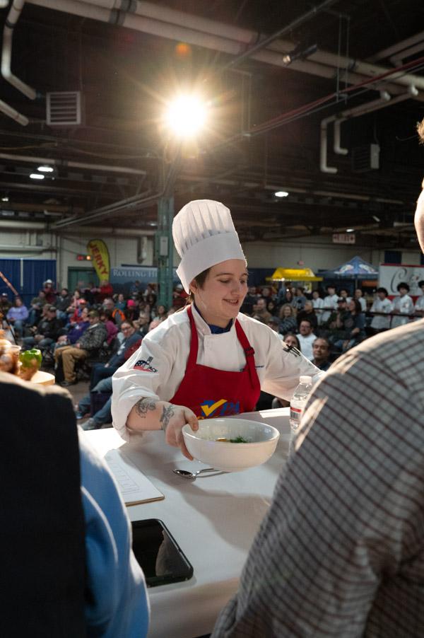 Chef presenting a plated dish to judges seated around a demonstration area.