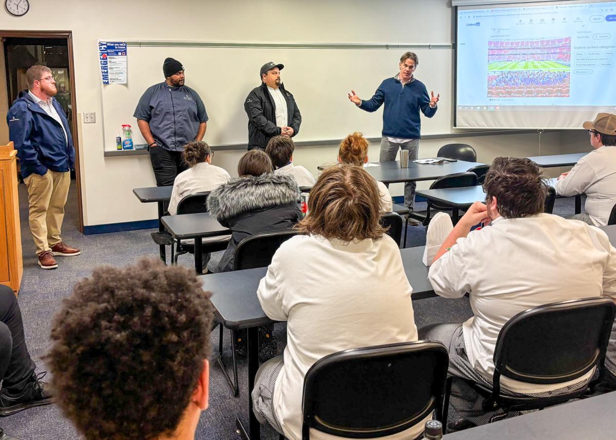 Four men stand in front of a classroom of students sitting at desks wearing chefs uniforms.