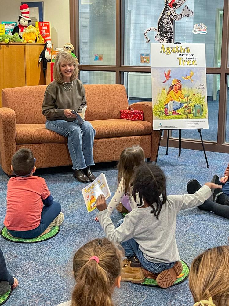A woman sits on a couch with a book in her hand while children sit on the floor looking at her.