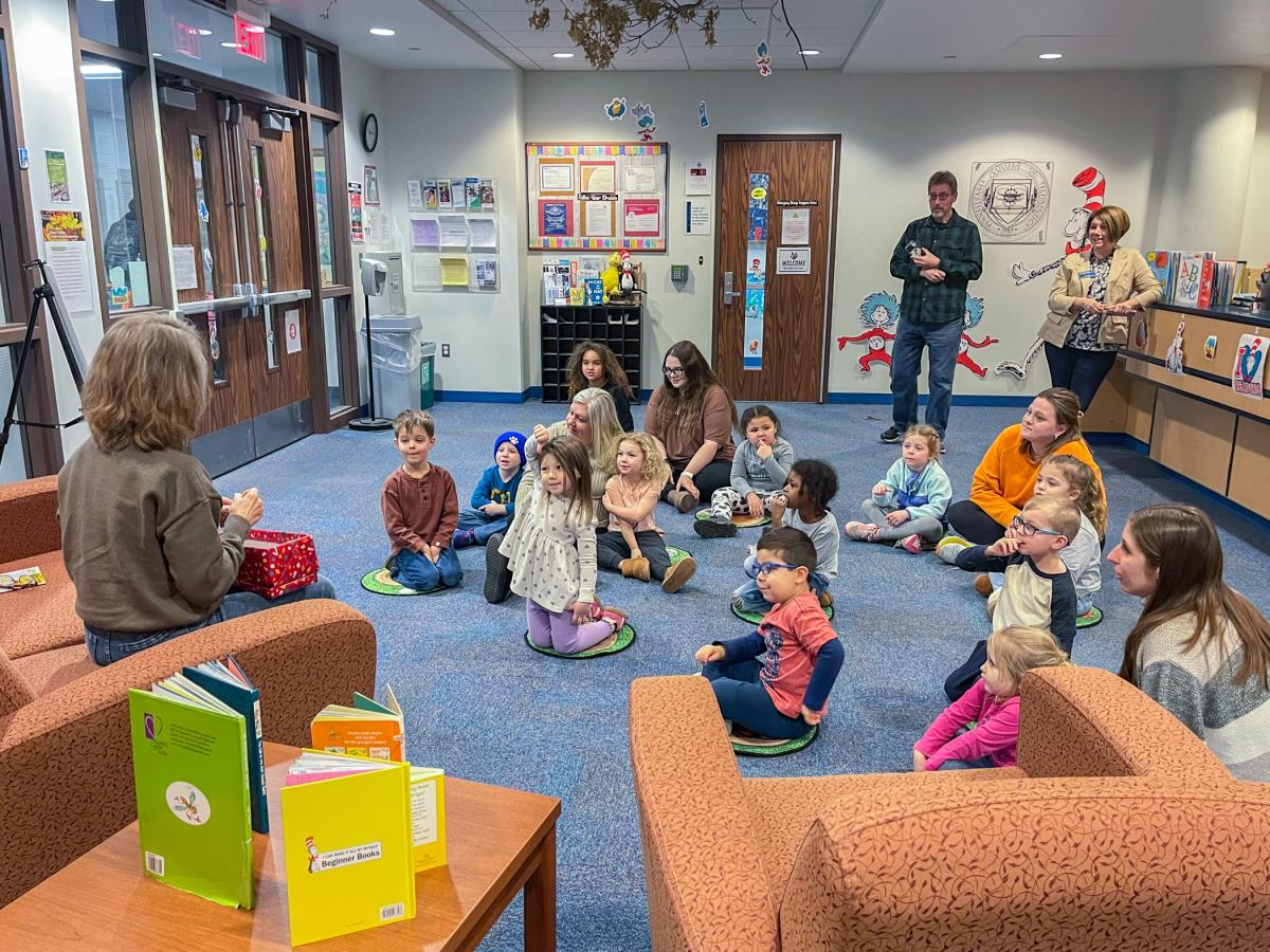 Preschool aged children sit on a carpeted classroom floor looking at a woman sitting on a couch. One child is raising her hand.
