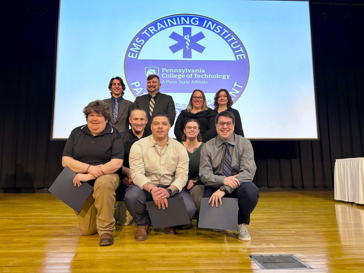 Nine people stand and kneel on Penn College's Klump Academic Center stage. Behind them is a large projection screen with a seal that reads "Pennsylvania College of Technology, EMS Training Institute, Paramedic Student."