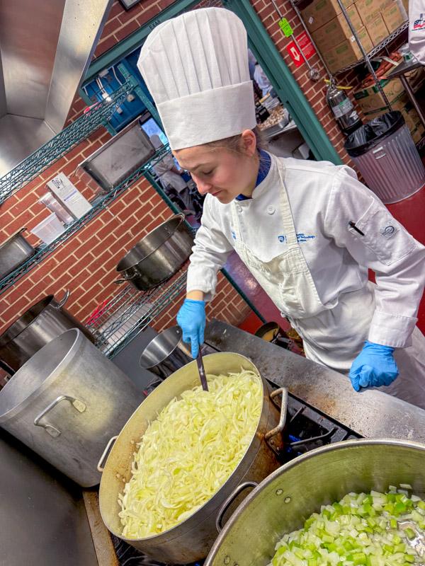 A student in a chef uniform stirs a large pot of vegetables.