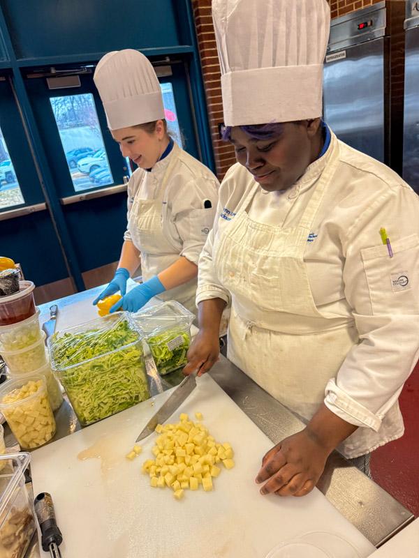 Two students in chef uniforms chop vegetables on cutting boards.