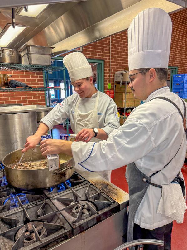Chefs work ove a large pot on a commercial stove.