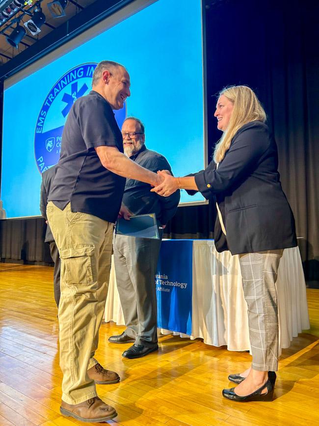 A man shakes a woman's hand. They are standing on a stage.