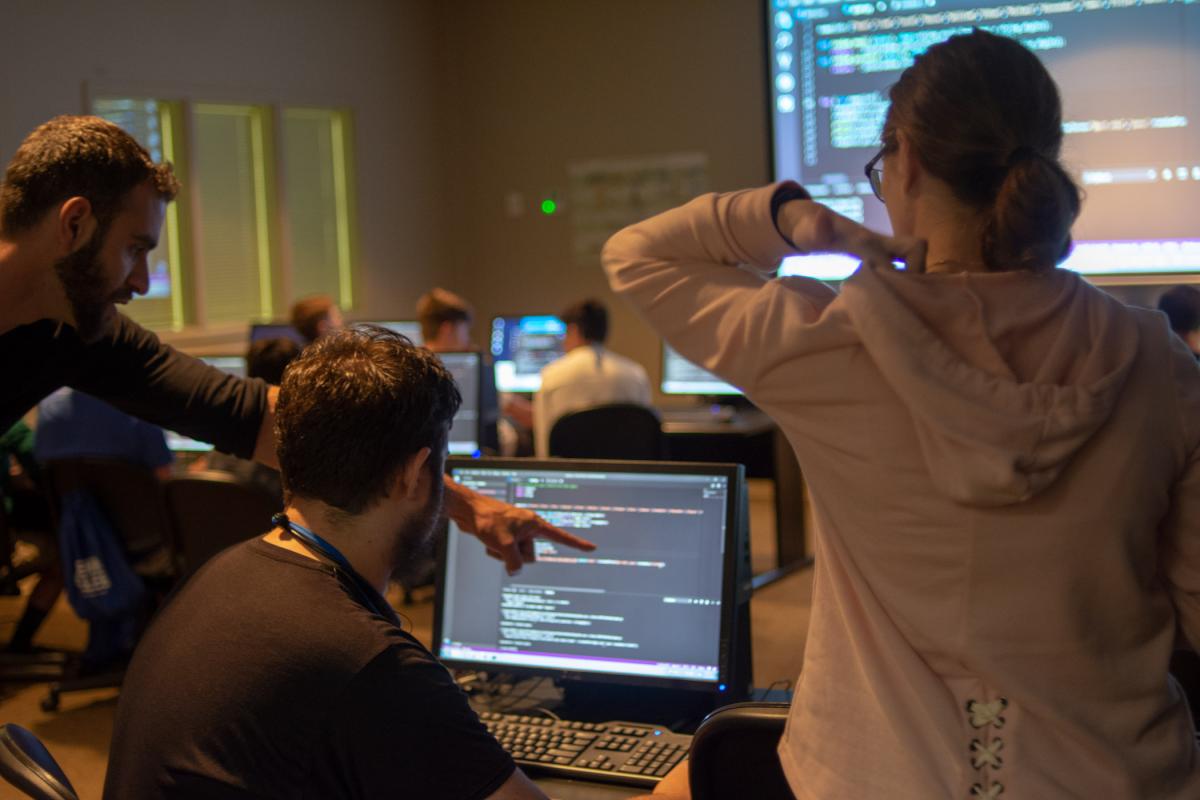 Three students review a projects displayed on a computer screen in a dimly lit lab.