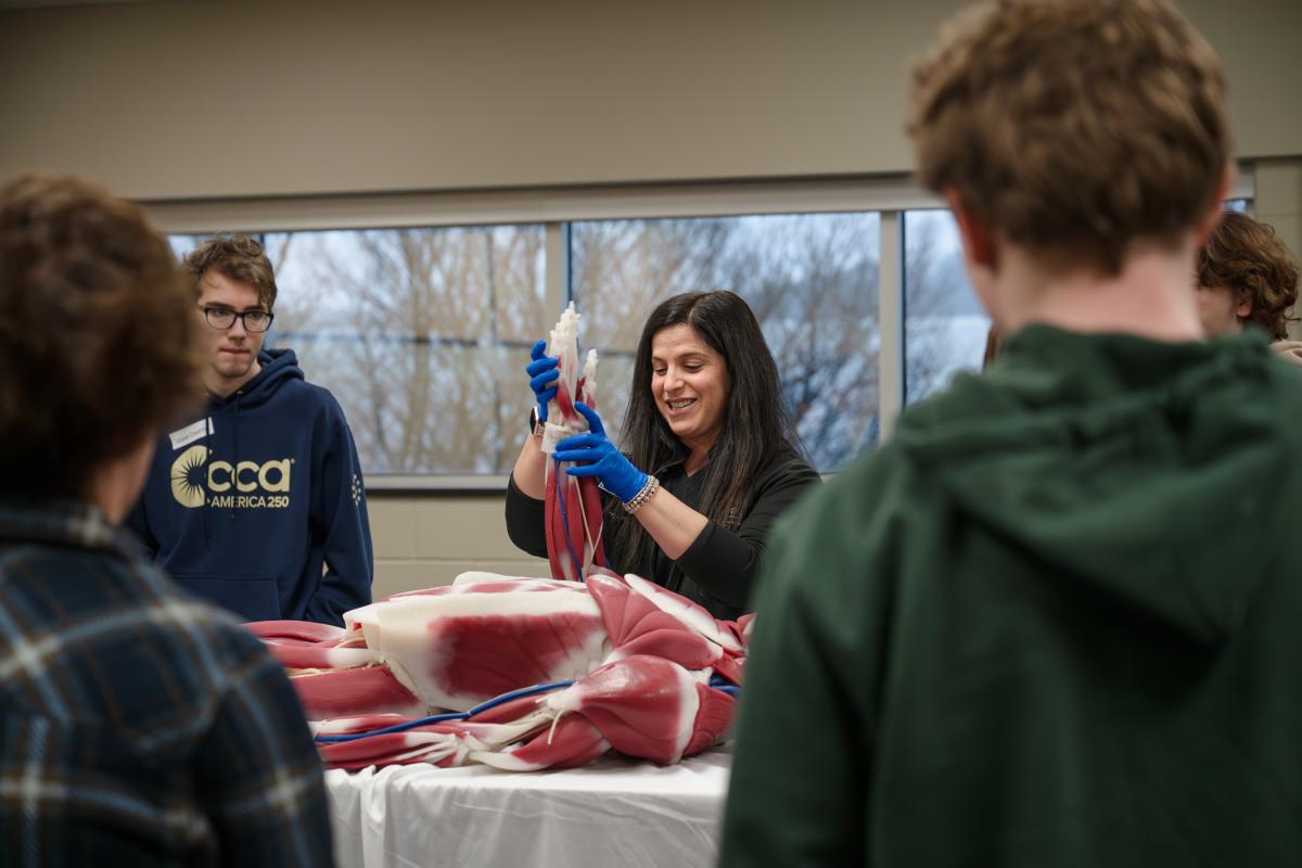 Surrounded by students, a woman holds up the arm of a specialized medical manikin.