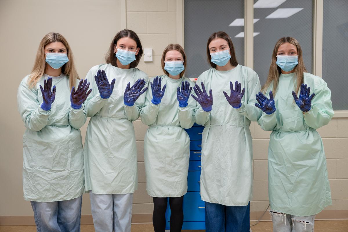Five high school students wearing surgical garb show the palms of their gloved hands.