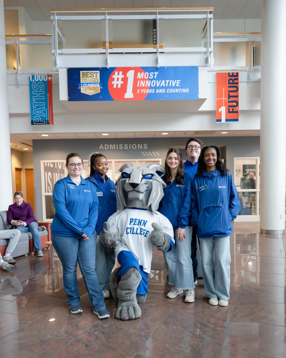 Students wearing blue shirts gather around Penn College's Wildcat mascot in the college's Davie Jane Gilmour Center.