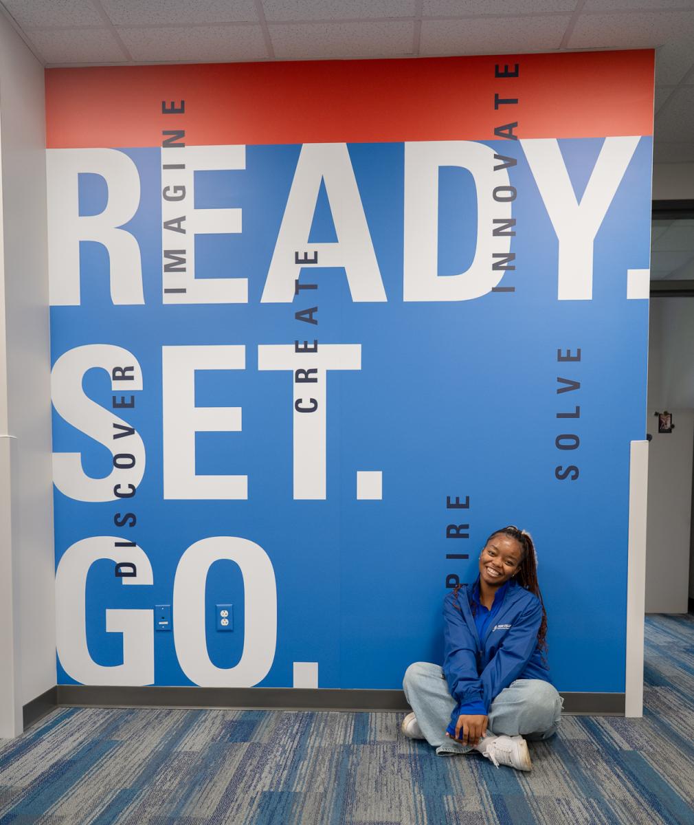 A student sits against a blue wall that reads, 'Ready Set Go.'