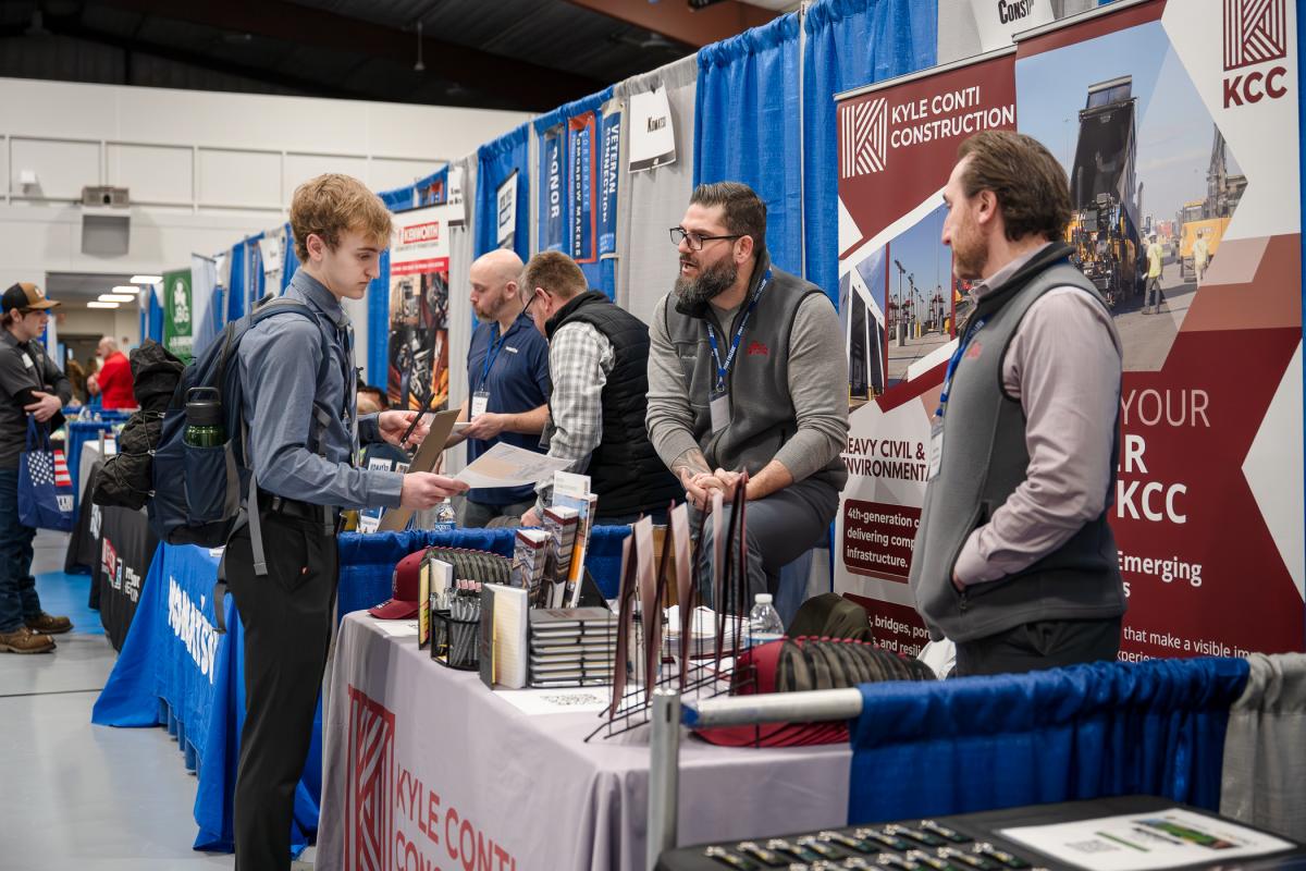 Men behind a Konti Construction table talk with a student who is wearing a dress shirt and tie.