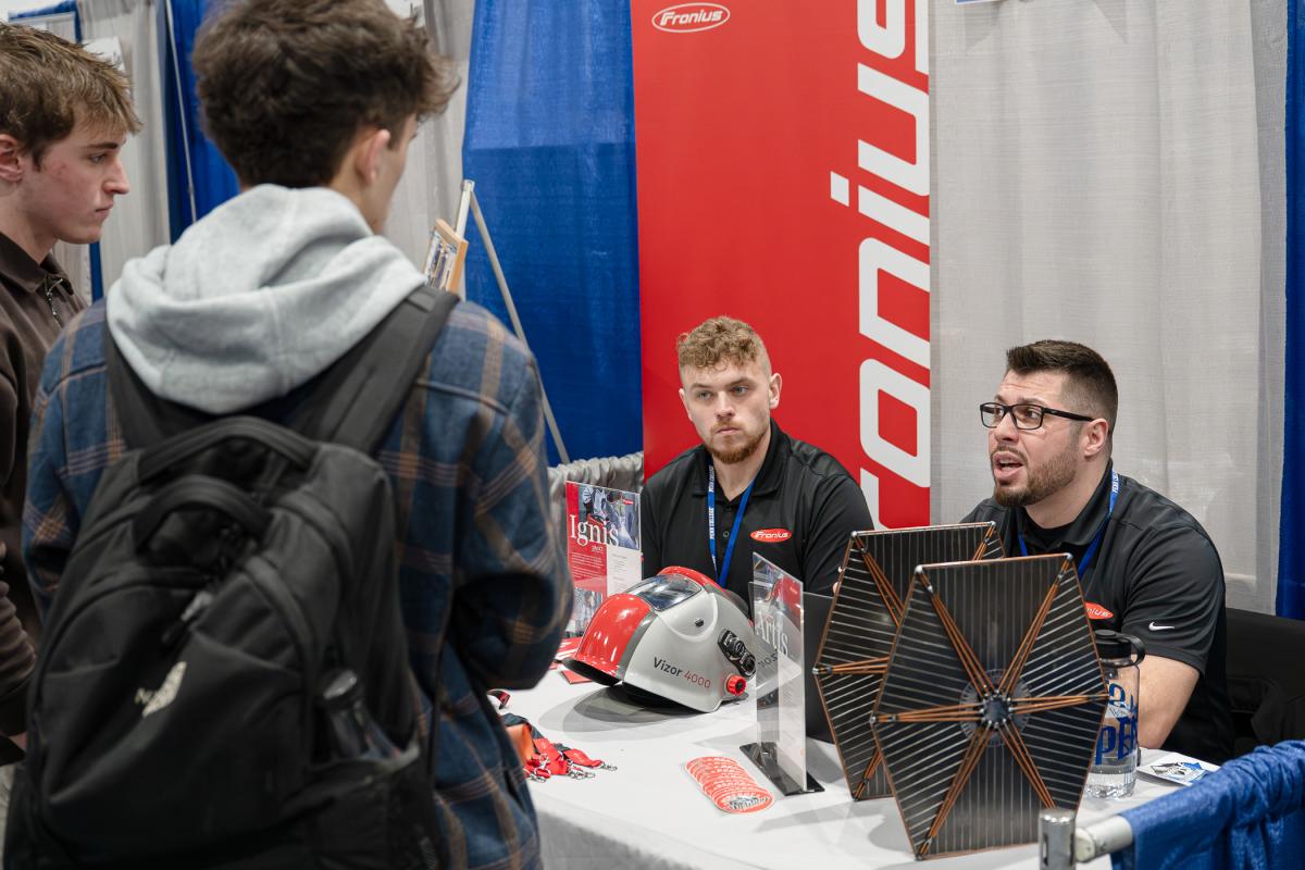 Students talk with two men wearing Fronius shirts and sitting behind a table of products and promotional materials from Fronius.