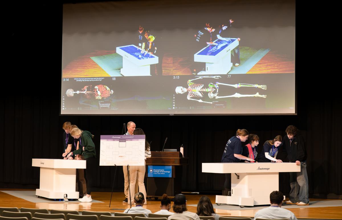 Two teams of high school students stand around their respective Anatomage tables on the Klump Academic Center stage while a large screen above projects their work for the audience.