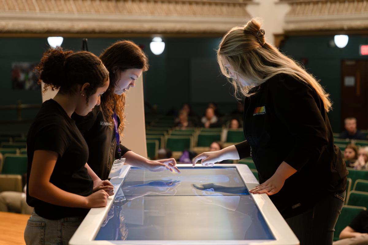On the stage in Penn College's Klump Academic Center Auditorium, three high school students stand around a large touchscreen table, tapping on a 3D anatomical image.