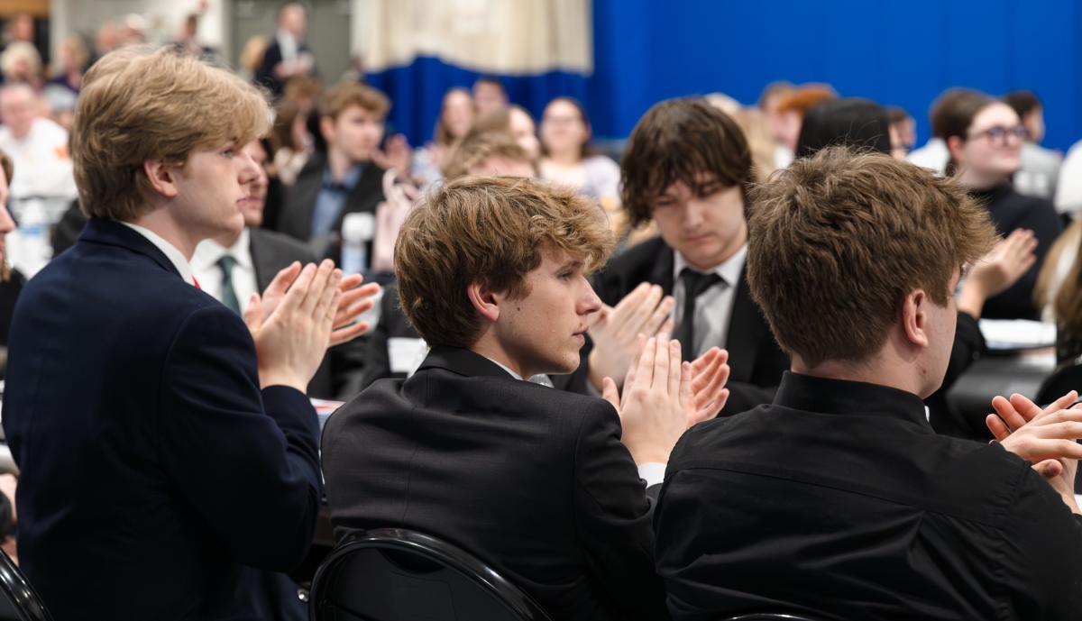 Students in black business suits clap while seated a table in a large room filled with other students sitting at tables.