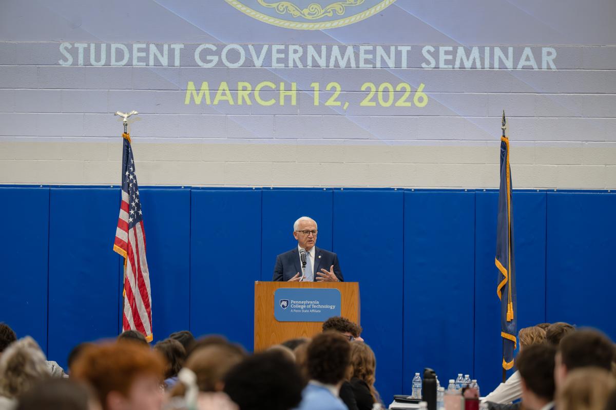 A man stands behind podium in a large room. A projection on the wall above him reads "Student Govermnet Seminar, March 12, 2026."