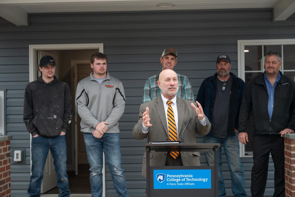A man speaks from a podium while Penn College students and faculty stand behind him on the porch of a house.