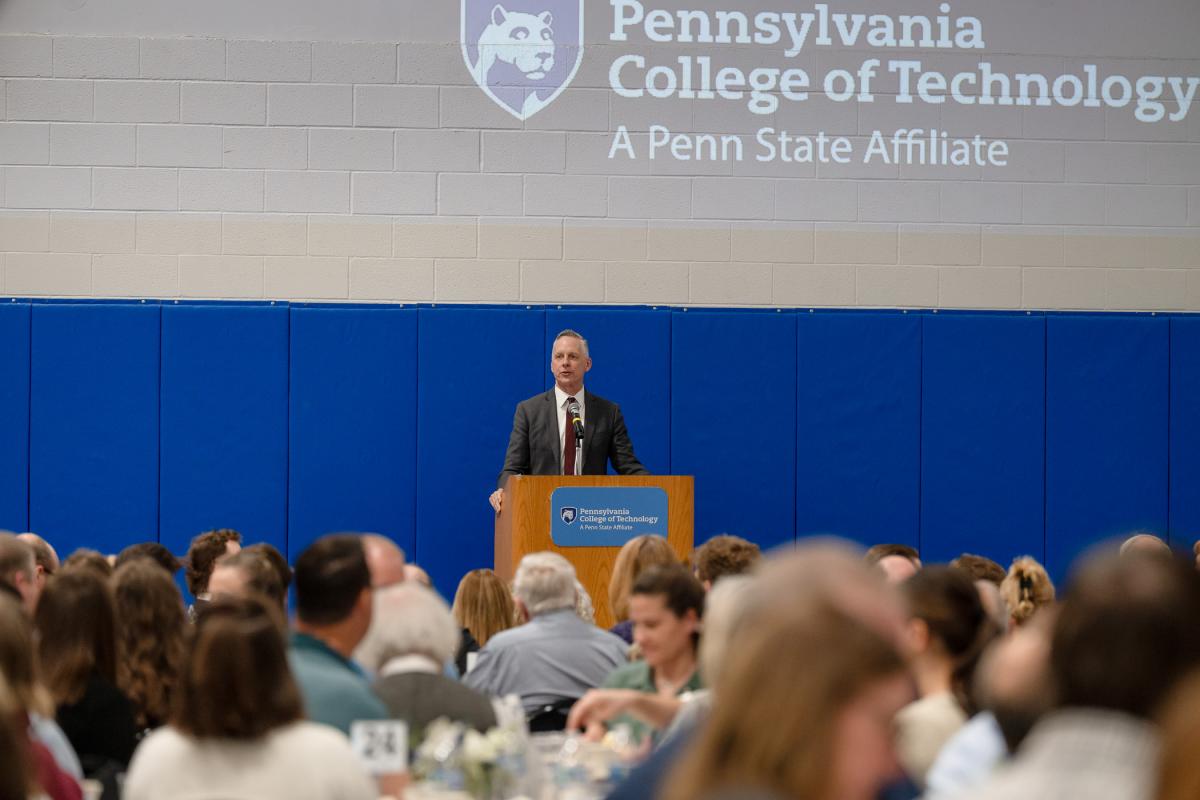 A man speaks from behind a podium in the college's Field House. In the foreground, people sit around tables.