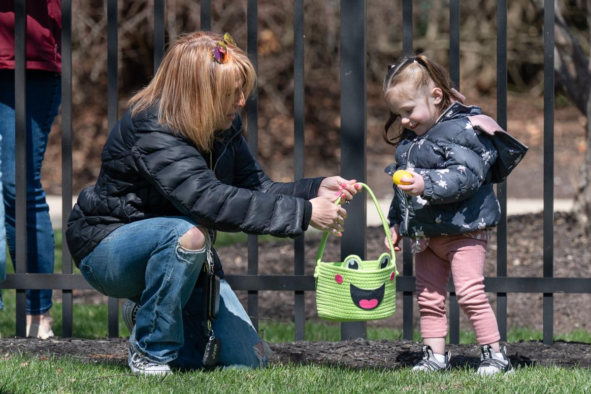 A woman kneels and holds a green Easter basket as a small smiling child places a plastic egg into it.