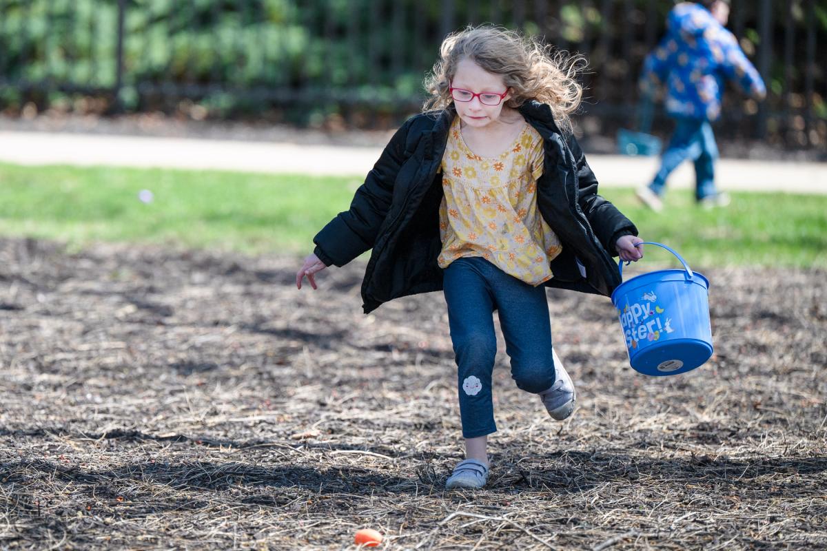 A child carrying a plastic bucket that says "Happy Easter" runs toward a plastic egg on the ground.