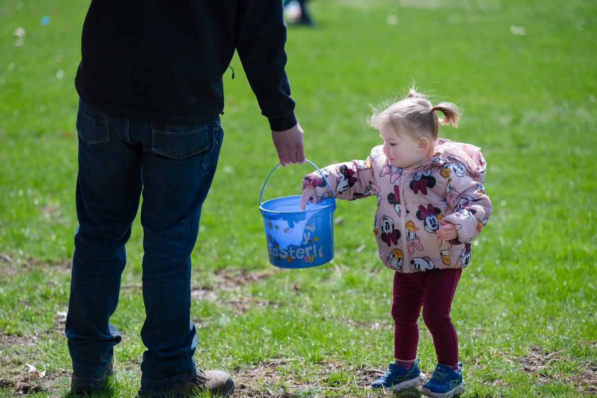 A small child places a plastic egg into a blue plastic bucket that says "Happy Easter" while an adult holds the bucket.