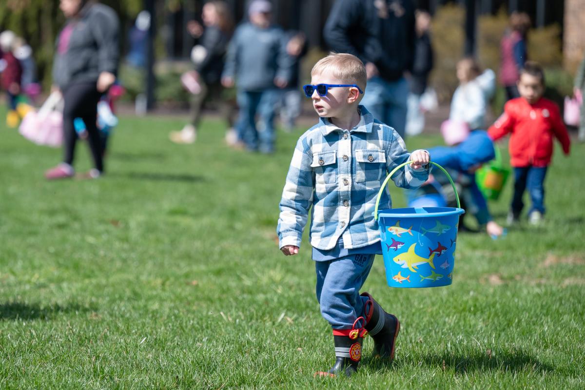 A child un sunglasses carries a bucket through a Penn College lawn, looking for eggs, as children in the background do the same.