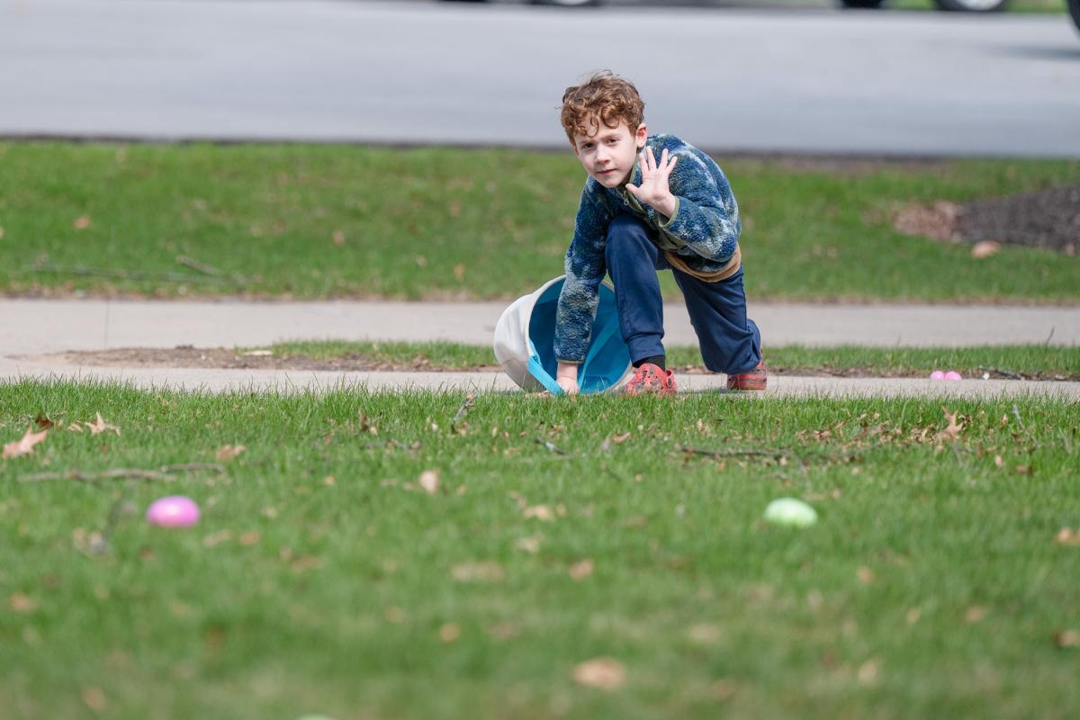 A child waves while knealing at the edge of a lawn, prepared to run toward plastic eggs.