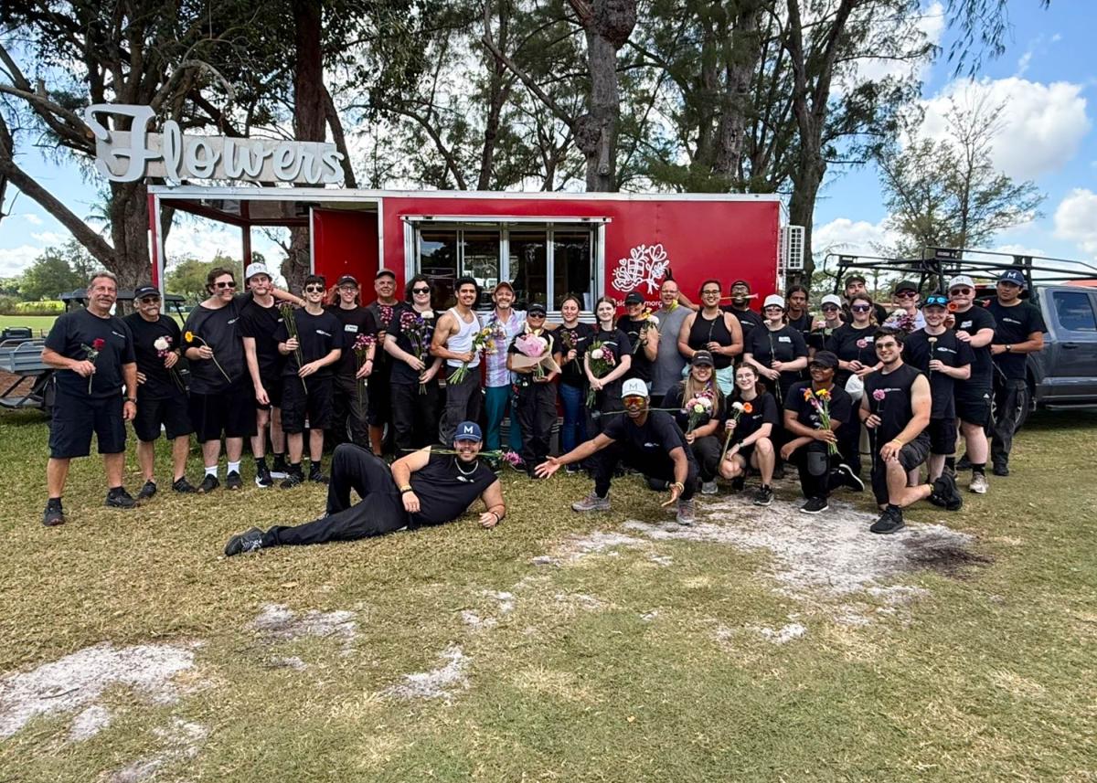 A group of people in black T-shirts and shorts gather in front of a flower vendor's trailer holding flowers.
