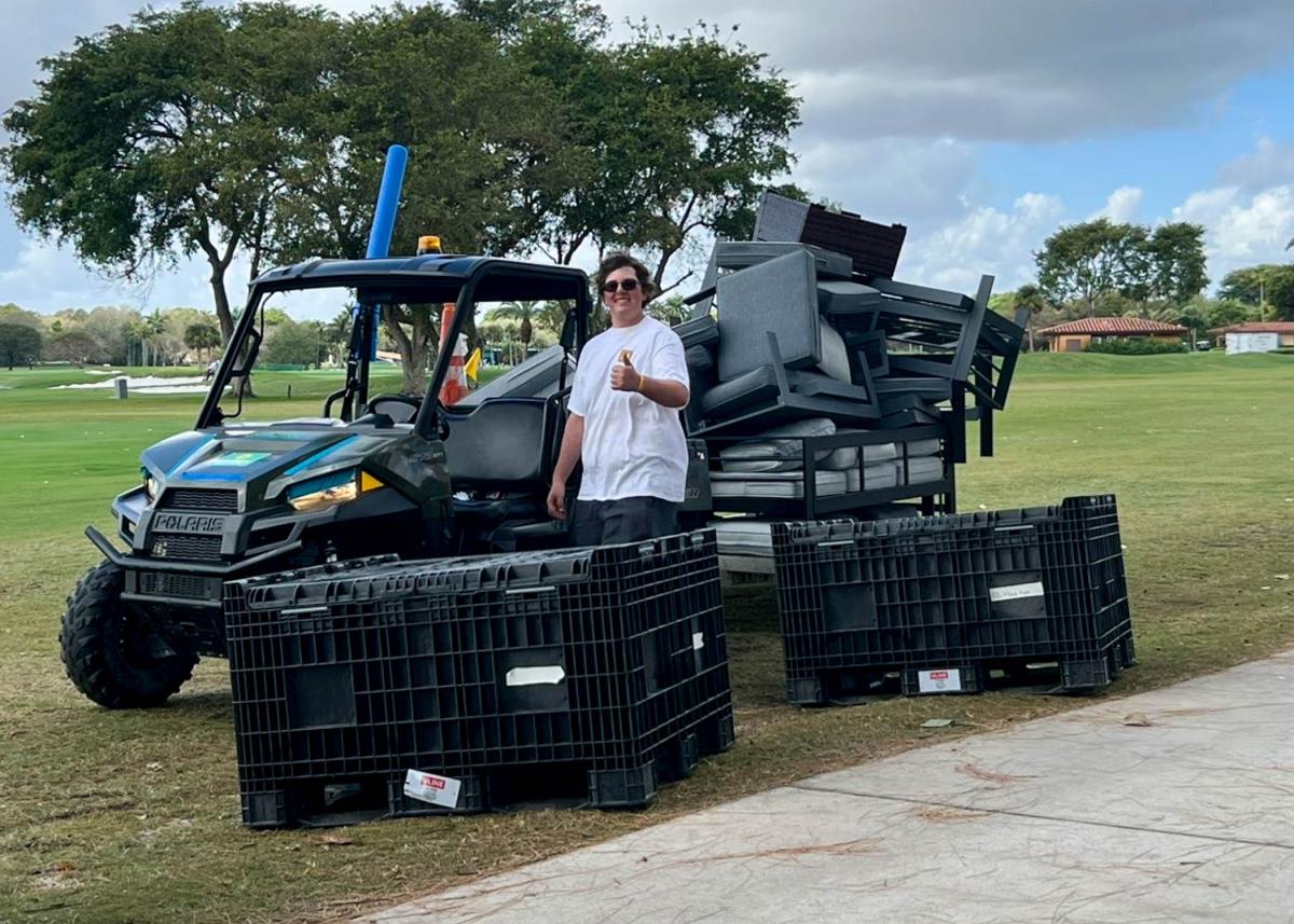 A student gives thumbs up as he stands next to a side-by-side vehicle that is pulling a cartload of padded chairs.