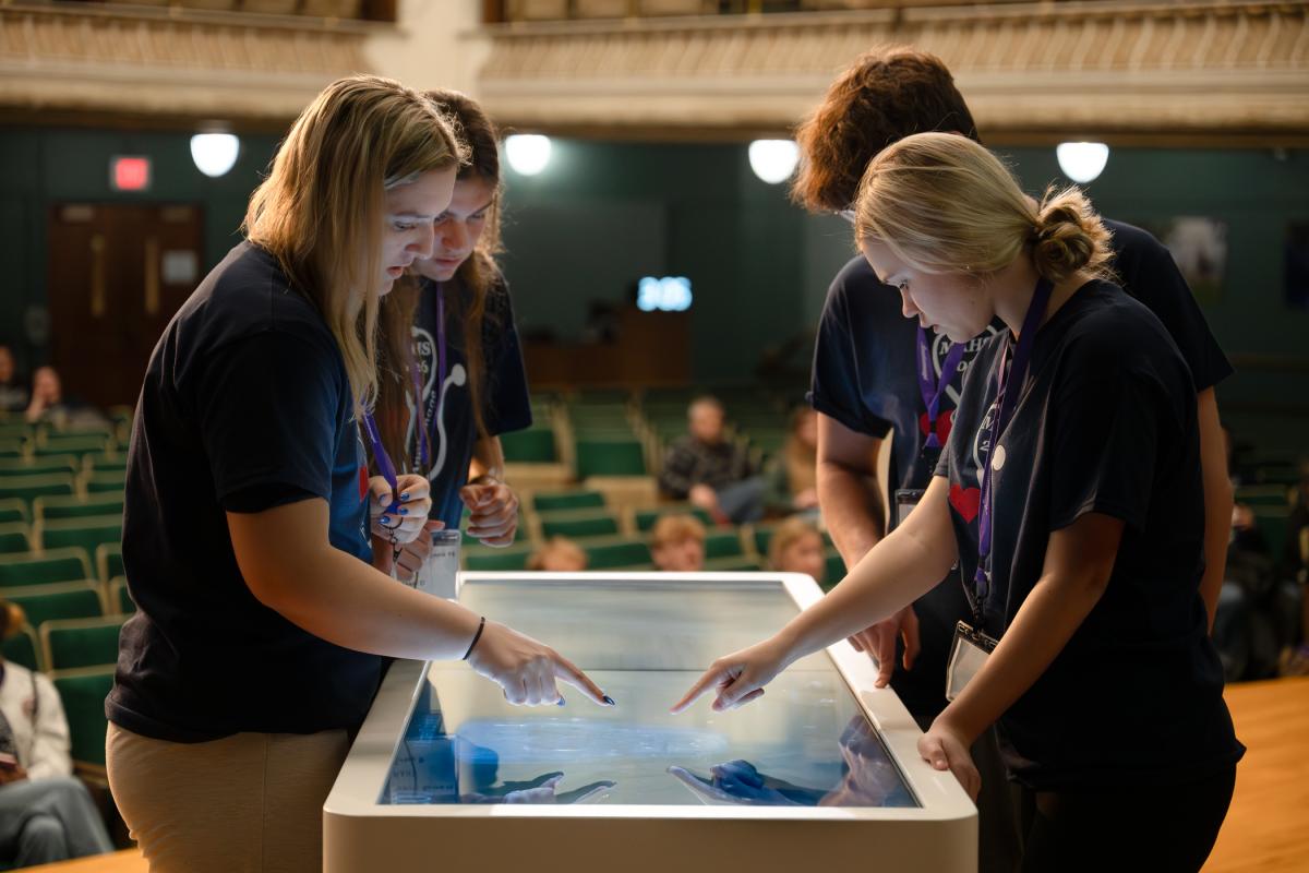 On the stage in Penn College's Klump Academic Center Auditorium, four high school students stand around a large touchscreen table, tapping on a 3D anatomical image.