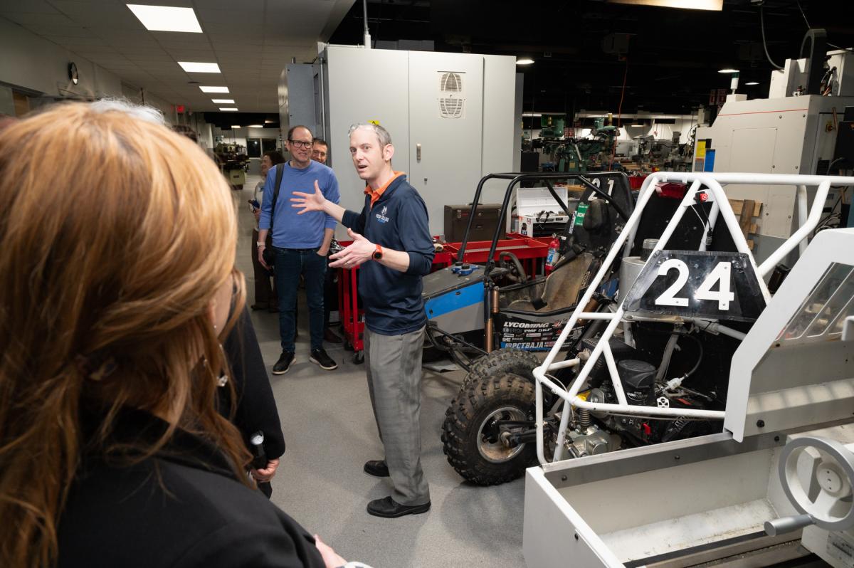A person standing in front of the frame of an off-road vehicle speaks to a group. They are surrounded by manufacturing equipment.